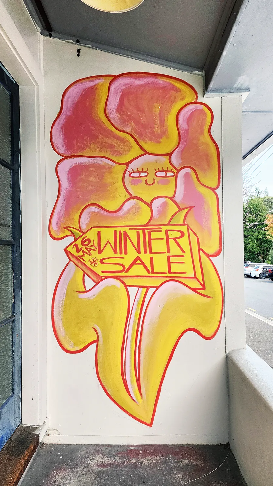 Happiness House mural - a smiling flower figure holding a Winter Sale sign, painted on the shop wall