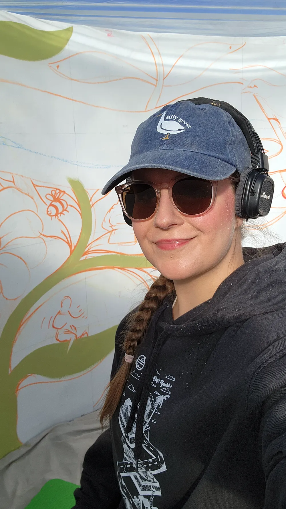 Kasia Hebda in a cap and headphones, smiling in front of the half-painted Onwards in Art! wall